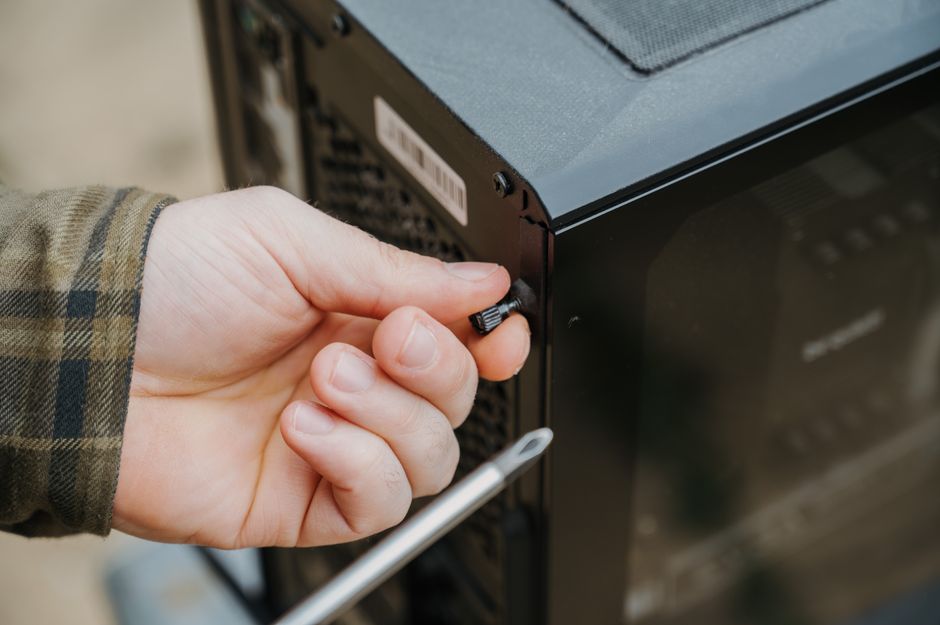 A close-up view of a person's hand adjusting a small screw on the rear panel of a black, modern garden machinery or electronic device, with a screwdriver resting nearby. The individual is wearing a pl